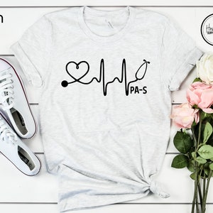 K&ouml;nnte beinhalten: Ein hellgraues T-Shirt mit einer schwarzen Grafik eines Herzens mit einem Stethoskop und einer Herzschlaglinie. Der Text "PA-S" befindet sich unter der Grafik.