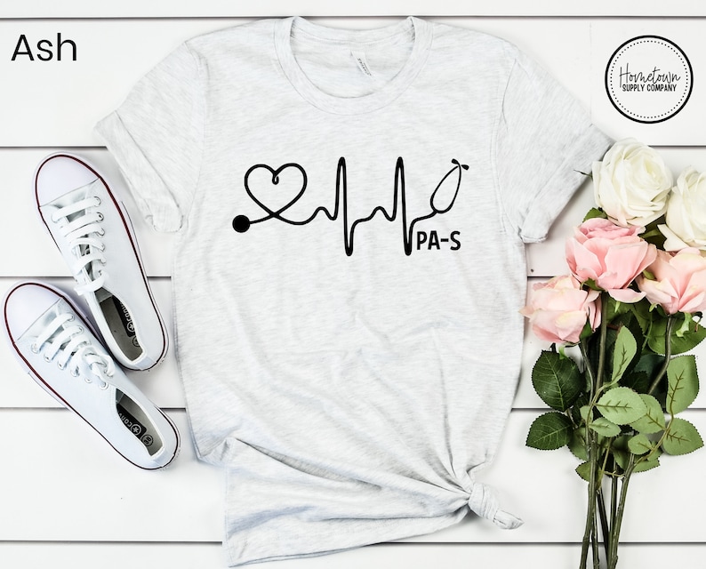 K&ouml;nnte beinhalten: Ein hellgraues T-Shirt mit einer schwarzen Grafik eines Herzens mit einem Stethoskop und einer Herzschlaglinie. Der Text "PA-S" befindet sich unter der Grafik.