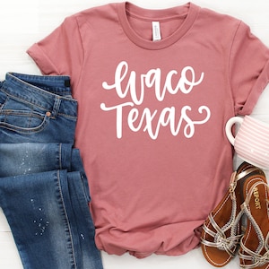 May include: A dusty rose t-shirt with the text "Waco Texas" in white script lettering. The shirt is paired with blue jeans and brown sandals.