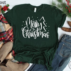 Puede incluir: Una camiseta verde con texto blanco que dice "Feliz Navidad" con un gr&aacute;fico de &aacute;rbol de Navidad estilizado. La camiseta est&aacute; colocada sobre una superficie plana con una manta a cuadros, botas marrones y pantalones vaqueros azules.