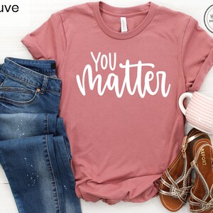 May include: Mauve t-shirt with the text "You Matter" in white lettering. The shirt is shown on a white surface with a pair of blue jeans, a pair of brown sandals, and a pink and white striped mug.