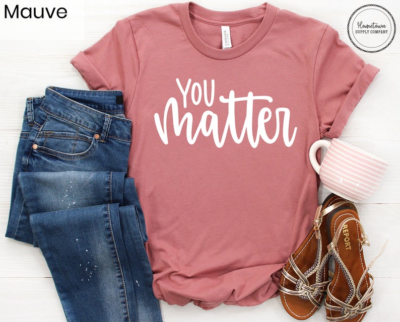 May include: Mauve t-shirt with the text "You Matter" in white lettering. The shirt is shown on a white surface with a pair of blue jeans, a pair of brown sandals, and a pink and white striped mug.