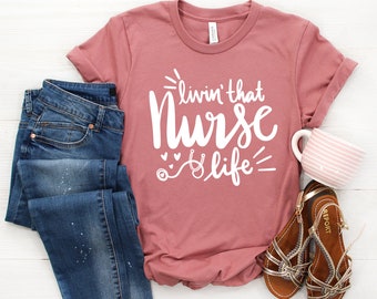 Puede incluir: Una camiseta rosa con el texto "livin' that Nurse life" en blanco. La camiseta está doblada y colocada sobre una superficie de madera blanca con un par de pantalones vaqueros azules, una taza y unas sandalias.