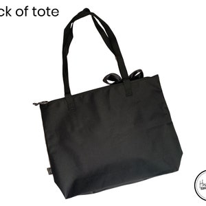 Może przedstawiać: Czarna torba typu tote z zamkiem błyskawicznym i kokardką na g&oacute;rze uchwyt&oacute;w. Torba ma napis "Hometown Supply Company" w prawym dolnym rogu.