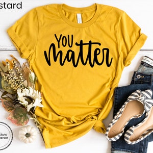 May include: A mustard yellow t-shirt with the text "You matter" in black script lettering. The shirt is laid flat on a white wooden surface with a bouquet of flowers, a small white pumpkin, and a pair of blue jeans and striped flats.
