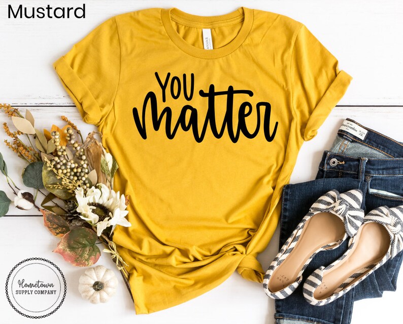 May include: A mustard yellow t-shirt with the text "You matter" in black script lettering. The shirt is laid flat on a white wooden surface with a bouquet of flowers, a small white pumpkin, and a pair of blue jeans and striped flats.