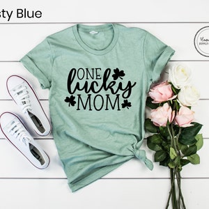 May include: A light green t-shirt with the text "One Lucky Mom" in black lettering and shamrock designs. The shirt is on a white wooden surface with a bouquet of pink and white roses.