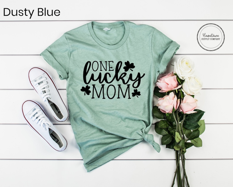 May include: A light green t-shirt with the text "One Lucky Mom" in black lettering and shamrock designs. The shirt is on a white wooden surface with a bouquet of pink and white roses.
