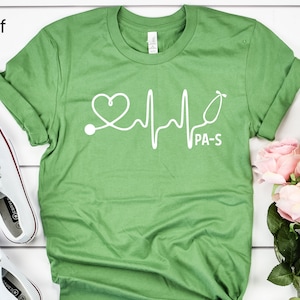 K&ouml;nnte beinhalten: Gr&uuml;nes T-Shirt mit einer wei&szlig;en Grafik eines Stethoskops und einem Herzschlag mit dem Text "PA-S".