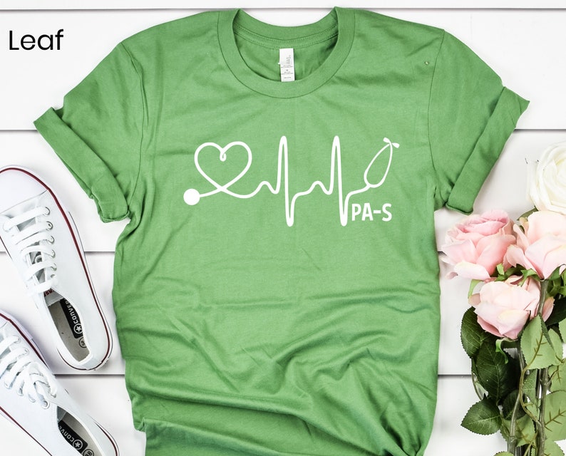 K&ouml;nnte beinhalten: Gr&uuml;nes T-Shirt mit einer wei&szlig;en Grafik eines Stethoskops und einem Herzschlag mit dem Text "PA-S".