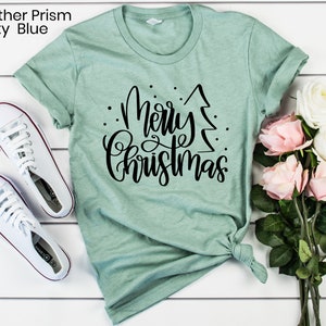 Puede incluir: Una camiseta verde claro con un dise&ntilde;o negro que dice "Feliz Navidad" con un &aacute;rbol de Navidad estilizado.