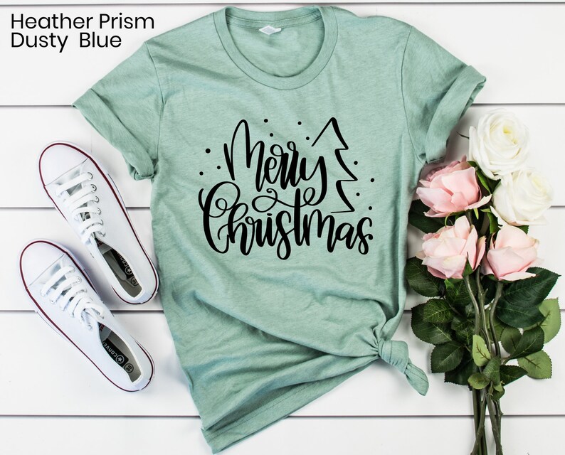 Puede incluir: Una camiseta verde claro con un dise&ntilde;o negro que dice "Feliz Navidad" con un &aacute;rbol de Navidad estilizado.