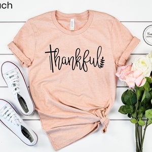 Puede incluir: Una camiseta de color melocot&oacute;n con la palabra "thankful" escrita en negro en cursiva, con un dise&ntilde;o de cruz y hoja. La camiseta est&aacute; colocada sobre una superficie de madera blanca con un par de zapatos de lona blancos y un ramo de rosas blancas y rosas.