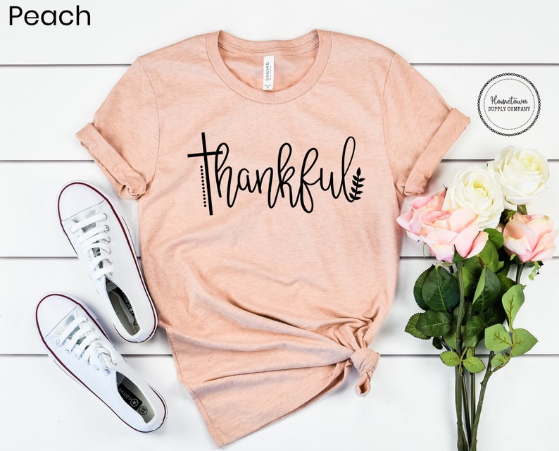 Puede incluir: Una camiseta de color melocot&oacute;n con la palabra "thankful" escrita en negro en cursiva, con un dise&ntilde;o de cruz y hoja. La camiseta est&aacute; colocada sobre una superficie de madera blanca con un par de zapatos de lona blancos y un ramo de rosas blancas y rosas.