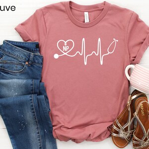 May include: Mauve t-shirt with a white graphic of a heart with the letters "NP" inside, a stethoscope, and a heartbeat line.