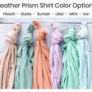K&ouml;nnte beinhalten: Sechs gefaltete T-Shirts in verschiedenen Pastellfarben: Pfirsich, staubiges Gr&uuml;n, Sonnenuntergangs-Orange, Flieder, Minzgr&uuml;n und Eisblau. Die T-Shirts sind in einer Reihe auf einer wei&szlig;en Holzoberfl&auml;che angeordnet.  Peach - Dusty - Sunset - Lilac - Mint - Ice
