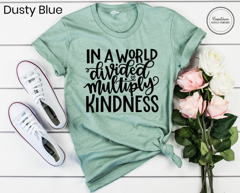 May include: A light green t-shirt with the text "In a world divided multiply kindness" in black lettering. The shirt is tied in a knot at the bottom and is lying on a white wooden surface. There are white sneakers and a bouquet of pink roses in the image.