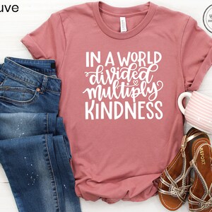May include: Mauve t-shirt with the text "In a world divided multiply kindness" in white lettering. The shirt is laid flat on a white surface with a pair of blue jeans, a mug, and sandals.