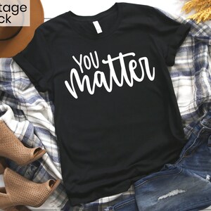 May include: Black t-shirt with white text that reads "You matter". The shirt is laid flat on a white surface with a plaid blanket, a brown hat, and blue jeans.