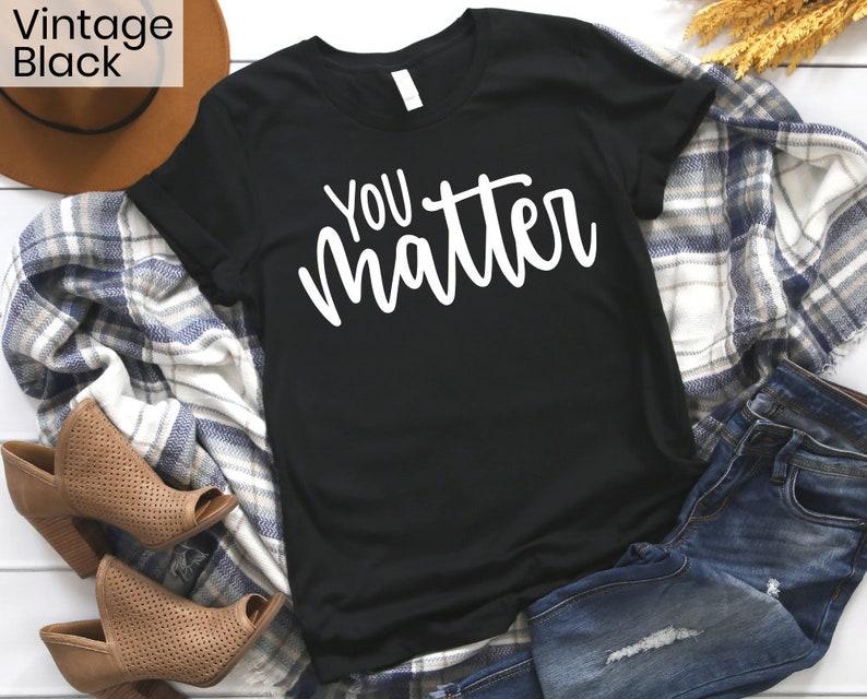 May include: Black t-shirt with white text that reads "You matter". The shirt is laid flat on a white surface with a plaid blanket, a brown hat, and blue jeans.