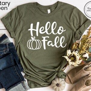 May include: A green t-shirt with the text "Hello Fall" in white lettering and a white pumpkin graphic. The shirt is folded and displayed on a white wooden surface with a pair of blue jeans, a small white pumpkin, and a bouquet of fall-colored flowers.