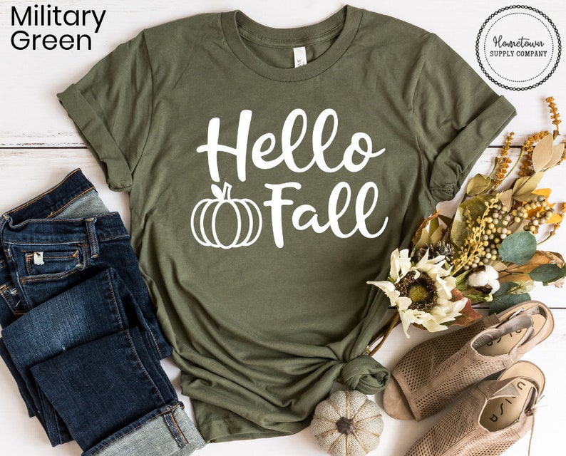 May include: A green t-shirt with the text "Hello Fall" in white lettering and a white pumpkin graphic. The shirt is folded and displayed on a white wooden surface with a pair of blue jeans, a small white pumpkin, and a bouquet of fall-colored flowers.