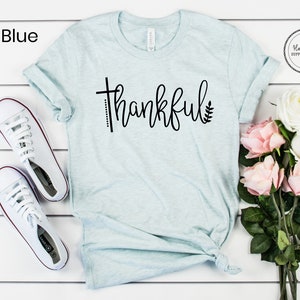Puede incluir: Una camiseta azul claro con la palabra "thankful" escrita en cursiva negra. La palabra est&aacute; acentuada con una peque&ntilde;a cruz y una hoja. La camiseta est&aacute; colocada sobre una superficie blanca con un par de zapatillas blancas y un ramo de rosas rosas y blancas.