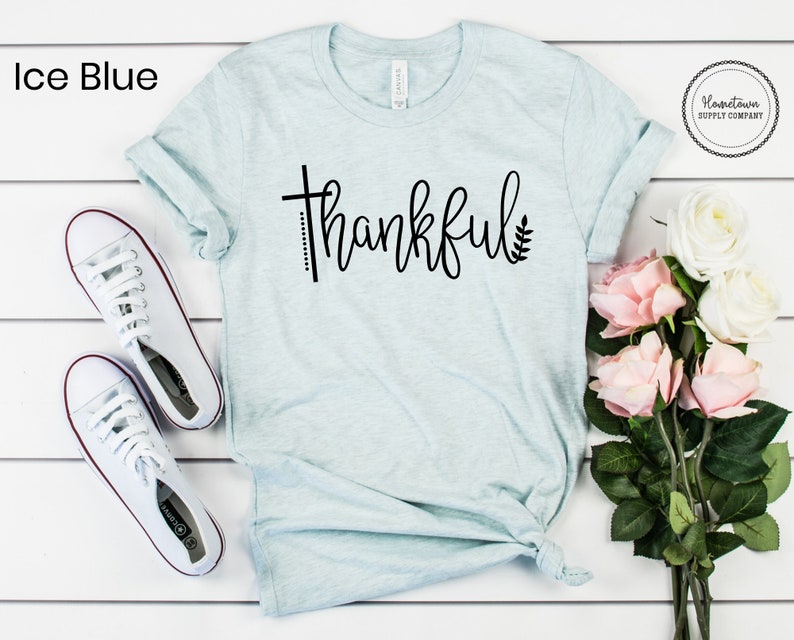 Puede incluir: Una camiseta azul claro con la palabra "thankful" escrita en cursiva negra. La palabra est&aacute; acentuada con una peque&ntilde;a cruz y una hoja. La camiseta est&aacute; colocada sobre una superficie blanca con un par de zapatillas blancas y un ramo de rosas rosas y blancas.