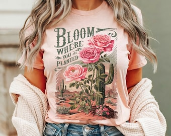 May include: A pink t-shirt with a graphic print of pink roses and a cactus. The text on the shirt reads "Bloom Where You Are Planted."