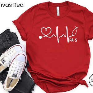 K&ouml;nnte beinhalten: Rotes T-Shirt mit einer wei&szlig;en Grafik eines Herzens, Stethoskops und einer EKG-Linie mit dem Text "PA-S".