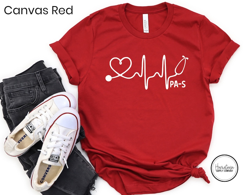 K&ouml;nnte beinhalten: Rotes T-Shirt mit einer wei&szlig;en Grafik eines Herzens, Stethoskops und einer EKG-Linie mit dem Text "PA-S".