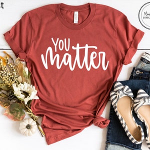 May include: A rust-colored t-shirt with the text "You matter" in white lettering. The shirt is laid flat on a white surface with a pair of striped flats and blue jeans.
