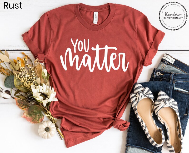 May include: A rust-colored t-shirt with the text "You matter" in white lettering. The shirt is laid flat on a white surface with a pair of striped flats and blue jeans.