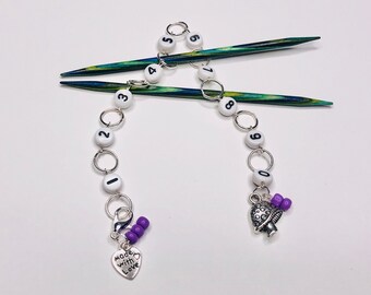 May include: A white bead stitch counter with black numbers 1 to 10, attached to two wooden knitting needles with a silver heart charm that says "Made with Love" and a silver mushroom charm with purple beads.