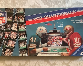 May include: A blue and red box for the NFL VCR Quarterback Game. The box features a photo of two football players, one in a blue and white uniform and the other in a red and white uniform. The box also features the text "Winner of 33 Emmys" and "VHS".