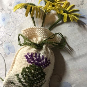 May include: A white knitted bag with a green and purple thistle design. The bag is filled with yellow crocheted flowers.