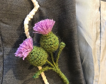 Puede incluir: Un broche de cardo tejido a ganchillo con dos cabezas de flor verdes texturizadas con puntas moradas vibrantes y deshilachadas, sujeto a un blazer gris oscuro con un borde tejido en blanco. El accesorio floral tiene tallos y hojas verdes.