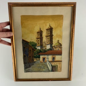 May include: Watercolor painting of a town square with two tall church towers. The buildings are painted in shades of brown, blue, and yellow. The painting is framed in a gold-colored frame.