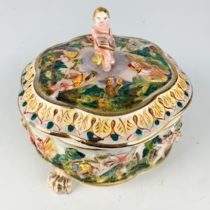 May include: A ceramic trinket box with a lid featuring a cherub figure holding a book. The box is decorated with colorful floral and figural designs, including cherubs, flowers, and leaves. The box has three feet and is painted in gold, green, yellow, red, and blue.
