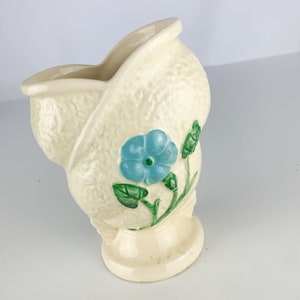 May include: A white ceramic vase with a textured surface and a blue and green floral design. The vase is shaped like a shell and has a wide opening.