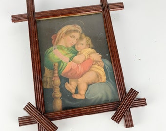 May include: A framed print of a painting depicting the Madonna and Child. The painting is in a dark wood frame with a decorative design. The frame is in the shape of a cross.