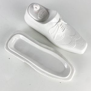 May include: A white ceramic butter dish shaped like a classic oxford shoe. The lid features detailed molded laces and brogue-style perforations, while the base is a rectangular tray with a decorative beaded edge. The interior is smooth and glossy.