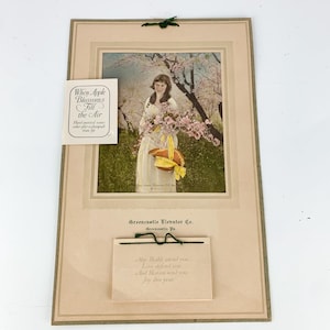 May include: Vintage calendar featuring a woman in a white dress holding a basket of pink blossoms under a blooming apple tree. The calendar is from the Greencastle Elevator Co. in Greencastle, Pennsylvania. The text reads: "When Apple Blossoms Fill the Air" and "May Health attend you, Live defend you, And Heaven send you Joy this year."