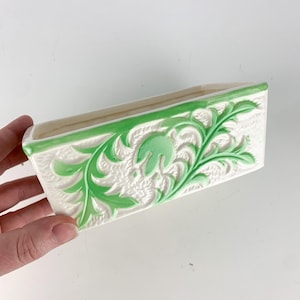 May include: A white ceramic planter with a green floral design. The planter is rectangular and has a slightly raised rim.