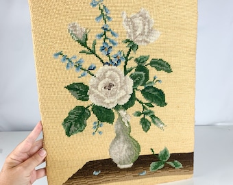 May include: A vintage needlepoint wall hanging featuring a bouquet of white roses in a green vase. The roses are surrounded by green leaves and blue flowers. The background is a light yellow.