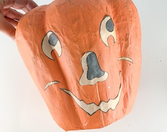 May include: A vintage paper mache pumpkin with a painted face. The pumpkin is orange with black and white details. The face has two large eyes, a small nose, and a wide smile.