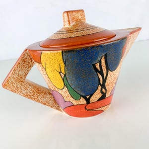 May include: A colorful ceramic teapot with a lid. The teapot features a speckled orange handle and body, with a lid and spout in a solid orange hue. The body is decorated with a vibrant abstract design in yellow, blue, green, and purple.