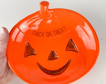 May include: Orange ceramic pumpkin-shaped dish with a glossy finish. The dish has a carved jack-o'-lantern face with a wide smile and the words "Trick or Treat" written on the top.