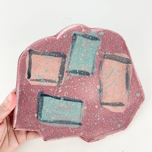 May include: A pink ceramic plate with an irregular shape and a speckled surface. The plate is decorated with four geometric shapes in black outlines, filled with pink and teal colors.