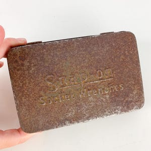 May include: A rectangular, rusty metal box with the embossed text "Snap-on Socket Wrenches." The box has a hinged lid and shows signs of age and wear, with a mottled, brown surface.